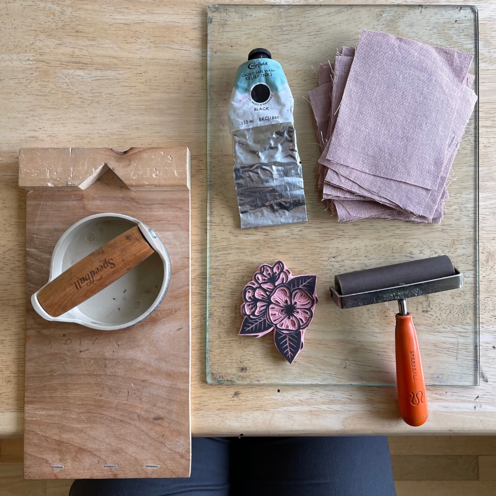 patch printing materials on table, to include a hand press, ink, glass plate, brayer, fabric, carved block, and bench hook.